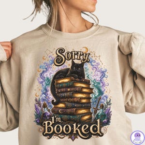 K&ouml;nnte beinhalten: Ein beigefarbenes Sweatshirt mit Rundhalsausschnitt und einem Grafikdruck einer schwarzen Katze, die auf einem Stapel alter B&uuml;cher sitzt. Das Design zeigt wirbelnde violette und blaue kosmische Wolken, Zaubertrankflaschen, ein Schwert und den Text Sorry, I'm BOOKED.