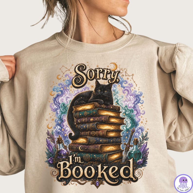 K&ouml;nnte beinhalten: Ein beigefarbenes Sweatshirt mit Rundhalsausschnitt und einem Grafikdruck einer schwarzen Katze, die auf einem Stapel alter B&uuml;cher sitzt. Das Design zeigt wirbelnde violette und blaue kosmische Wolken, Zaubertrankflaschen, ein Schwert und den Text Sorry, I'm BOOKED.