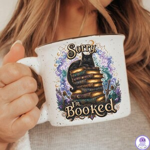 K&ouml;nnte beinhalten: Ein wei&szlig;er Keramikbecher mit gesprenkelter Oberfl&auml;che und einer Grafik einer schwarzen Katze, die auf einem Stapel alter B&uuml;cher sitzt. Der Text lautet Sorry I'm BOOKED, umgeben von bunten wirbelnden Wolken, kleinen Mondsicheln und Zaubertrankflaschen.