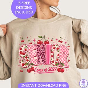 Op de afbeelding: Een beige sweater met ronde hals en een grafische print met het woord SENIOR in grote letters. De letters hebben roze ruitjes, luipaardprint en gestreepte patronen. Het ontwerp bevat rode kersen, groene bladeren en roze sterren.