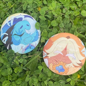 Pode incluir: Dois botões redondos com ilustrações de desenhos animados coloridos. Um botão apresenta uma criatura azul com um fundo branco e azul. O outro botão tem um personagem com cabelo laranja e amarelo e uma flor.