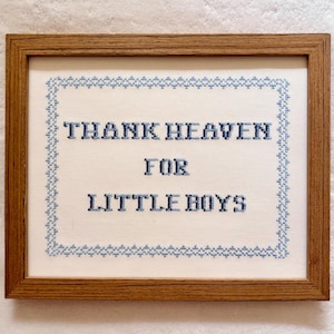 May include: A framed cross-stitch artwork with the text "THANK HEAVEN FOR LITTLE BOYS" in blue thread. The text is centered on a white background, surrounded by a blue border, and encased in a brown wooden frame.