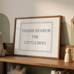 May include: A framed cross-stitch artwork with the text "THANK HEAVEN FOR LITTLE BOYS" in blue thread. The artwork is in a wooden frame and sits on a wooden shelf. The background is a light beige color.