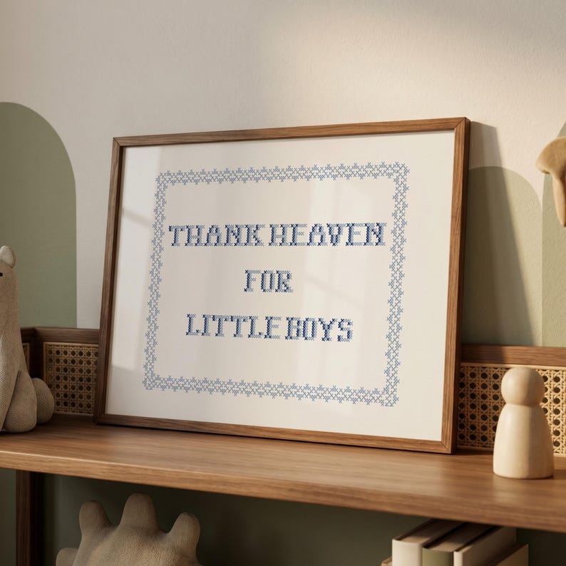 May include: A framed cross-stitch artwork with the text "THANK HEAVEN FOR LITTLE BOYS" in blue thread. The artwork is in a wooden frame and sits on a wooden shelf. The background is a light beige color.
