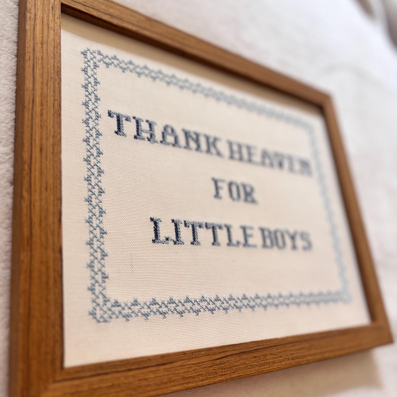 May include: A framed cross-stitch piece with the text "THANK HEAVEN FOR LITTLE BOYS" in dark blue thread. The text is centered on a cream-colored fabric, framed by a blue stitched border and a wooden frame.