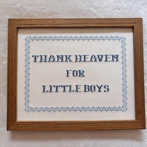 May include: A framed cross-stitch artwork with the words "THANK HEAVEN FOR LITTLE BOYS" in blue thread. The text is centered on a white fabric background, surrounded by a blue border, and encased in a brown wooden frame.