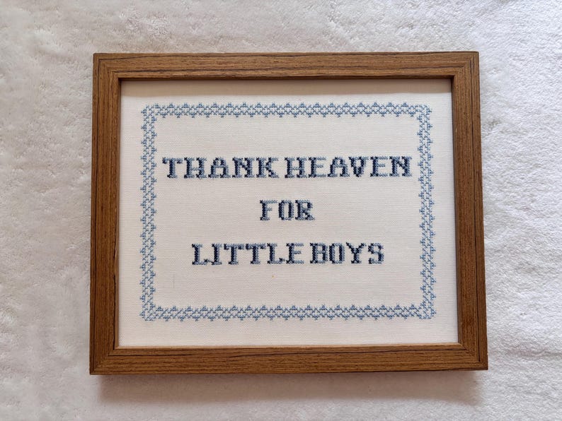 May include: A framed cross-stitch artwork with the words "THANK HEAVEN FOR LITTLE BOYS" in blue thread. The text is centered on a white fabric background, surrounded by a blue border, and encased in a brown wooden frame.
