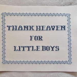 May include: A white fabric cross-stitch with the words "THANK HEAVEN FOR LITTLE BOYS" in dark blue thread. The text is centered within a blue stitched border. The fabric has a slightly textured appearance.