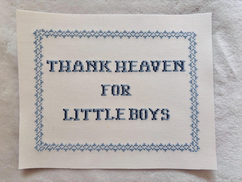 May include: A white fabric cross-stitch with the words "THANK HEAVEN FOR LITTLE BOYS" in dark blue thread. The text is centered within a blue stitched border. The fabric has a slightly textured appearance.