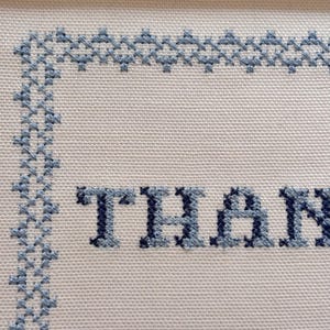 May include: Close-up of a cross-stitch design on white fabric. The word "THAN" is stitched in blue thread. A blue border frames the text, with a decorative pattern along the top and left edges.
