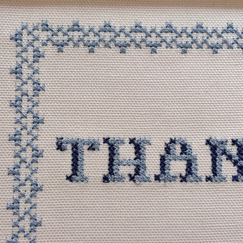 May include: Close-up of a cross-stitch design on white fabric. The word "THAN" is stitched in blue thread. A blue border frames the text, with a decorative pattern along the top and left edges.