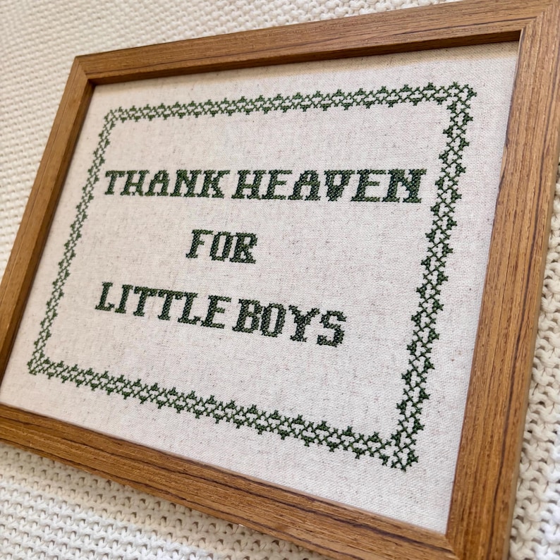 May include: Cross-stitch artwork in a wooden frame. The text "THANK HEAVEN FOR LITTLE BOYS" is stitched in dark green thread on a light-colored fabric. A decorative green border surrounds the text.