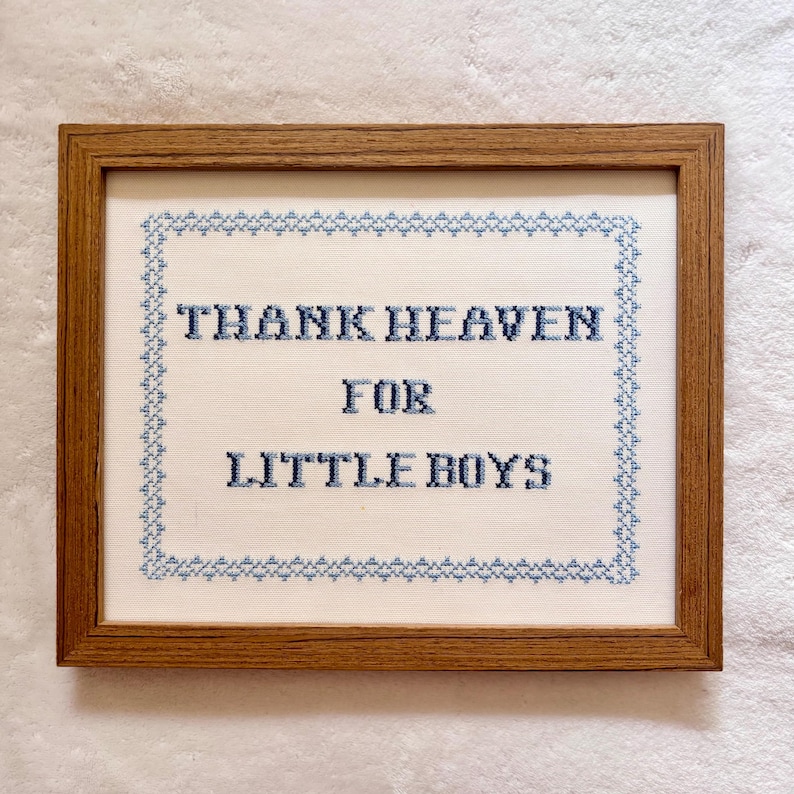 May include: A framed cross-stitch artwork with the text "THANK HEAVEN FOR LITTLE BOYS" in blue thread. The text is centered on a white background, surrounded by a blue border, and encased in a brown wooden frame.