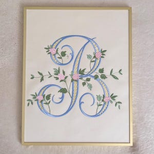 May include: Embroidered artwork featuring a large, ornate letter "B" in light blue, adorned with pink roses and green leaves. The artwork is framed in a gold-colored frame, set against a cream-colored background.