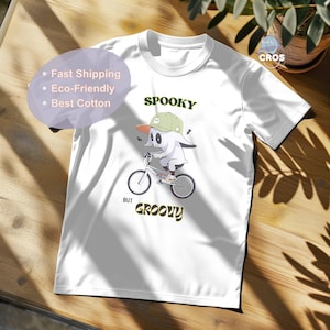 Puede incluir: Camiseta blanca con un gráfico de un fantasma montando una bicicleta. El fantasma lleva una gorra de béisbol verde y blanca. El texto "Spooky but Groovy" está impreso debajo del fantasma.