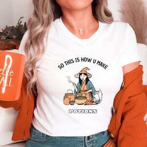 May include: White t-shirt with a graphic of a witch making potions with a cat. The text on the shirt reads "So this is how u make potions".