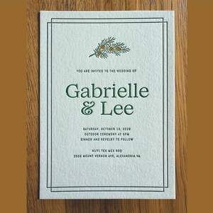 May include: A rectangular white wedding invitation card with a double-line border, featuring a green pine branch illustration, the text "You are invited to the wedding of Gabrielle & Lee", date, time, and location details in dark green ink.