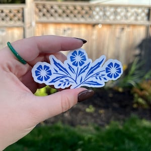 May include: A white sticker with a blue floral design. The sticker features three stylized flowers and several leaves. The design is in a classic blue and white color scheme, reminiscent of traditional porcelain patterns.