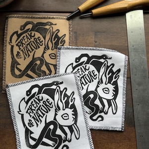 May include: Square fabric patches with a black and white graphic design. The design features a rabbit with snakes and the text "Freak of Nature". One patch is tan, and the others are white with black stitching.