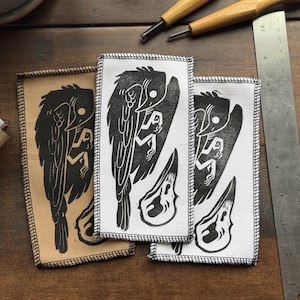 May include: Three fabric patches with a black and white bird and figure design. The patches are rectangular with stitched edges. One patch is tan, and two are white. A metal ruler and carving tools are also visible.