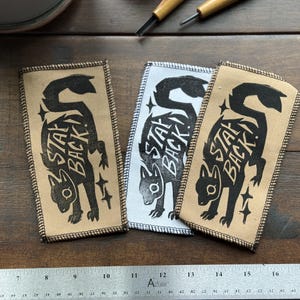 May include: Three rectangular patches with a black squirrel illustration and the words "STAY BACK!" printed on them. The patches are in shades of tan and white, with black stitching around the edges. A ruler is at the bottom.