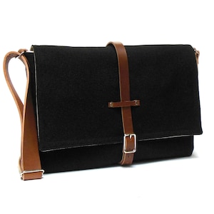 Può includere: Una borsa a tracolla in tela nera con una tracolla e una fibbia in pelle marrone. La borsa ha una chiusura a lembo e una sola tracolla.