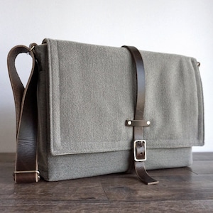 Può includere: Una borsa a tracolla in tela grigia con una tracolla e una fibbia in pelle marrone. La borsa ha una chiusura a lembo e un ampio scomparto principale.