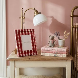 May include: A red and white gingham picture frame with the text "7 MONKIES" stands on a wooden table. A brass lamp with a white shade and a stack of pink books are also on the table. A small stuffed mouse with a pink scarf is on top of the books.