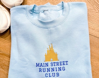 Könnte beinhalten: Ein hellblaues Sweatshirt mit Rundhalsausschnitt und einer goldenen Schloss-Stickerei über dem blauen Schriftzug MAIN STREET RUNNING CLUB. Das Kleidungsstück hat einen gerippten Kragen und lange Ärmel, dargestellt in flacher Ansicht.