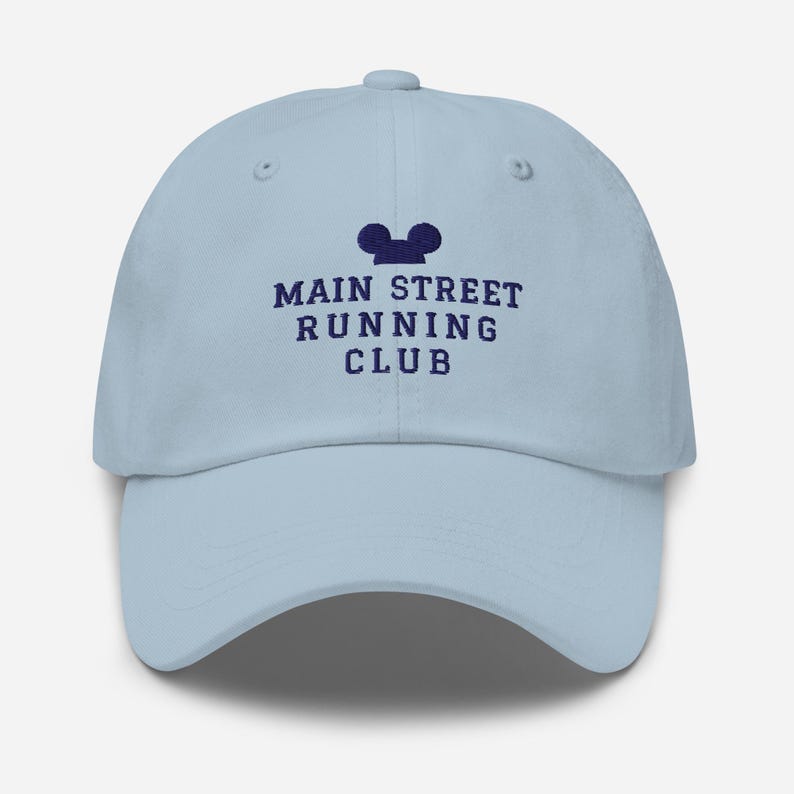 K&ouml;nnte beinhalten: Eine hellblaue Baseballkappe mit dunkelblauer Stickerei auf der Vorderseite. Das Design zeigt eine Silhouette von Mausohren &uuml;ber dem Text MAIN STREET RUNNING CLUB in einer fetten Schriftart. Die Kappe hat einen gebogenen Schirm.