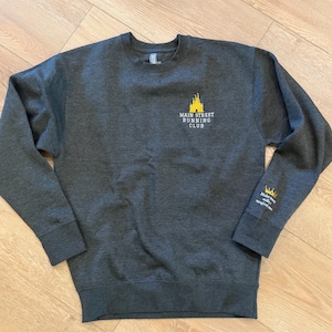 May include: A dark charcoal gray crewneck sweatshirt featuring a yellow castle graphic and the text "MAIN STREET RUNNING CLUB" on the chest. The left sleeve cuff displays a small yellow crown icon and the text "Make every mile magical."