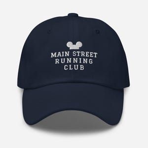 K&ouml;nnte beinhalten: Eine marineblaue Baseballkappe mit wei&szlig;er Stickerei auf der Vorderseite. Das Design zeigt ein stilisiertes Mausohren-Symbol &uuml;ber dem Text MAIN STREET RUNNING CLUB, der in drei Zeilen in einer klaren, serifenlosen Schriftart angeordnet ist.