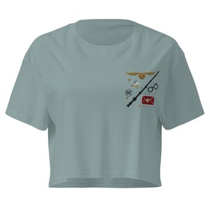 May include: A sage green cropped t-shirt featuring a small embroidered design on the left chest. The embroidery includes a golden snitch, a black wand, round glasses, a red envelope with a gold seal, and the number 9 3/4 in a black circle.