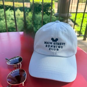 May include: A light blue baseball cap with the embroidered text MAIN STREET RUNNING CLUB and a mouse ear icon in dark blue, accompanied by a pair of dark-tinted sunglasses with thin silver frames resting on a red surface.