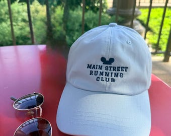 Puede incluir: Una gorra de béisbol azul claro con el texto bordado MAIN STREET RUNNING CLUB y un icono de orejas de ratón en azul oscuro, acompañada de unas gafas de sol oscuras con montura plateada fina sobre una superficie roja.