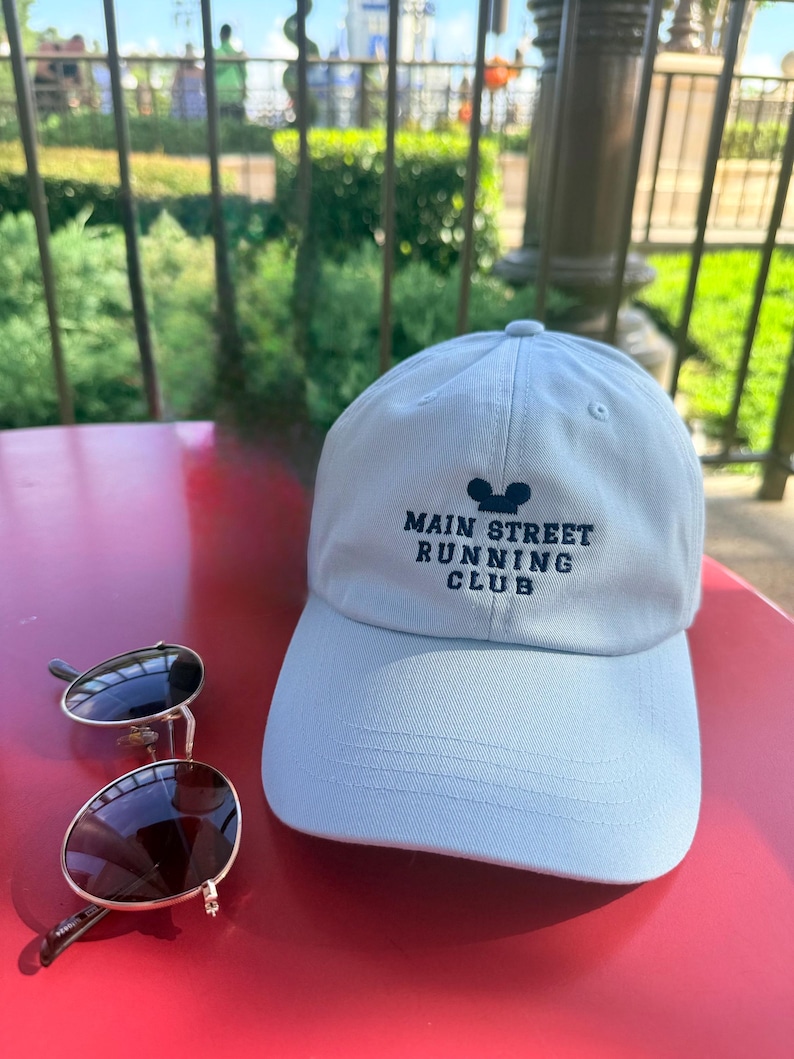 K&ouml;nnte beinhalten: Eine hellblaue Baseballkappe mit dem gestickten Text MAIN STREET RUNNING CLUB und einem Mausohren-Symbol in Dunkelblau, begleitet von einer dunkel get&ouml;nten Sonnenbrille mit d&uuml;nnem silbernen Rahmen auf einer roten Oberfl&auml;che.
