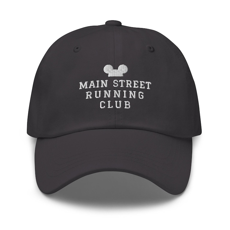 K&ouml;nnte beinhalten: Eine anthrazitfarbene Baseballkappe mit wei&szlig;em Sticktext, der MAIN STREET RUNNING CLUB lautet, platziert unter einem kleinen, wei&szlig; gestickten Symbol in Form einer stilisierten Mauskopf-Silhouette auf der vorderen Kappe.