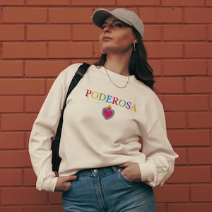 May include: A white long-sleeved sweatshirt with the word "PODEROSA" printed in rainbow colors. A red heart with a flame on top is centered below the word. The sweatshirt has a black bag strap on the shoulder and the words "CULTURA ES RESISTENCIA" printed on the left sleeve.