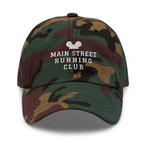 K&ouml;nnte beinhalten: Eine Baseballkappe mit Camouflage-Muster in Gr&uuml;n, Braun und Schwarz. Auf der Vorderseite befindet sich ein wei&szlig;er Stickschriftzug mit dem Text MAIN STREET RUNNING CLUB, der unter einem kleinen wei&szlig;en Stick-Symbol von Mausohren platziert ist.