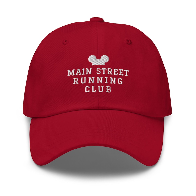 K&ouml;nnte beinhalten: Eine rote Baseballkappe mit wei&szlig;er Stickerei auf der Vorderseite. Das Design zeigt ein stilisiertes Mausohren-Symbol &uuml;ber dem Text MAIN STREET RUNNING CLUB in einer Serifenschrift. Die Kappe hat einen gebogenen Schirm und eine strukturierte Krone.