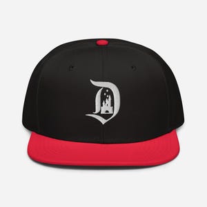May include: Black baseball cap with a red peak and a white embroidered "D" logo featuring a castle design. The cap has a flat peak and a red button on top. A classic and stylish accessory.