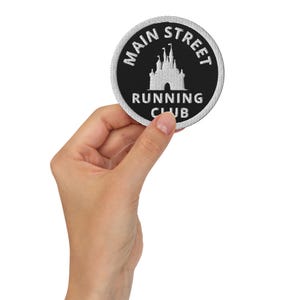 May include: A round black and white patch with the text "Main Street Running Club" and a castle graphic. The patch is held up by a hand.