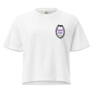 Può includere: T-shirt bianca con girocollo e maniche corte. La maglietta presenta una grafica viola e nera sul davanti con la scritta "Ghost Host" all'interno di una cornice ovale decorativa con un pipistrello in cima. Il testo "EST. 1999" è in basso.