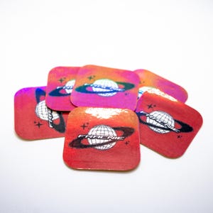 May include: A stack of red, pink, and orange holographic stickers with a black and white graphic of a planet with a ring around it. The text "FRACNIE" is printed on the sticker.