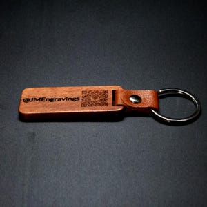May include: A wooden keychain with a brown leather strap and a silver key ring. The wooden part has the text "@JMEngravings" and a QR code engraved on it.