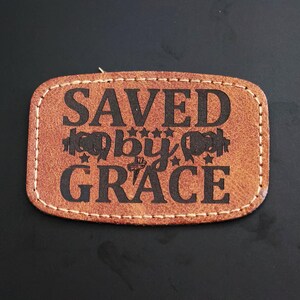 May include: A brown leather patch with the text "SAVED by GRACE" in black lettering. The text is surrounded by a decorative design of hearts and stars.