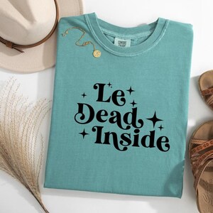 May include: A teal blue t-shirt with the text "Le Dead Inside" in black, retro-style font. The text is surrounded by small gold stars.