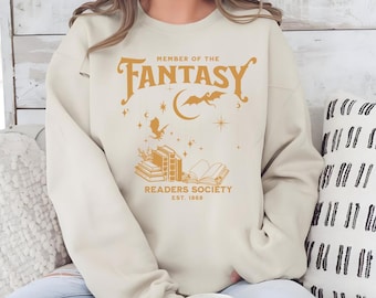 May include: A cream coloured sweatshirt with a gold graphic design that says "Member of the Fantasy Readers Society Est. 1868". The graphic includes a crescent moon, stars, and a dragon.