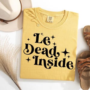 May include: A yellow t-shirt with the text "Le++ Dead++ + Inside" in black. The shirt has a distressed look and is made of a soft, comfortable fabric.