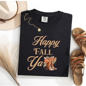 May include: A black short-sleeved t-shirt with the text Happy FALL YA in gold lettering. The graphic includes a pair of brown cowboy boots filled with orange and white flowers. A gold chain necklace rests on the shirt near the collar.
